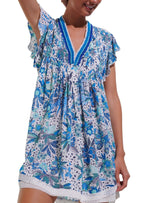 A woman wears a short, sleeveless Poupette St Barth Sasha Mini Dress adorned with blue and green floral patterns. The dress, embodying true beach style from Poupette St Barth, features a V-neckline and ruffled sleeves. Only the upper half of her body is visible.