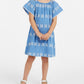 A young girl wearing the Mer Girls’ Mini Capucine Caftan with white embroidered detailing and white sandals, standing against a plain white background.
