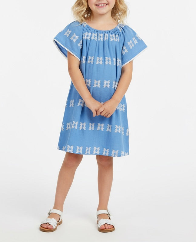 A young girl wearing the Mer Girls’ Mini Capucine Caftan with white embroidered detailing and white sandals, standing against a plain white background.