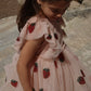 A young girl with long brown hair stands outdoors on stone wearing the Konges Slojd Girls' Yvonne Frill Dress, a voluminous light pink tulle dress adorned with embroidered strawberries.