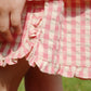 A close-up of a child’s hand resting on Konges Slojd Girls' Kim Frill Shorts in pink and white checkered seersucker fabric with ruffled edges, outdoors on a sunny day.