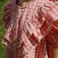 A girl stands outdoors in sunlight wearing the Konges Slojd Girls' Kim Frill Shirt, featuring pink and white checks, seersucker fabric, ruffled shoulders, and delicate frills.