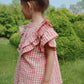 A young child with brown hair in a bun stands outdoors on grass, wearing Konges Slojd Girls' Kim Frill Shorts in pink and white gingham. Green bushes and trees form the background as she faces to the side.