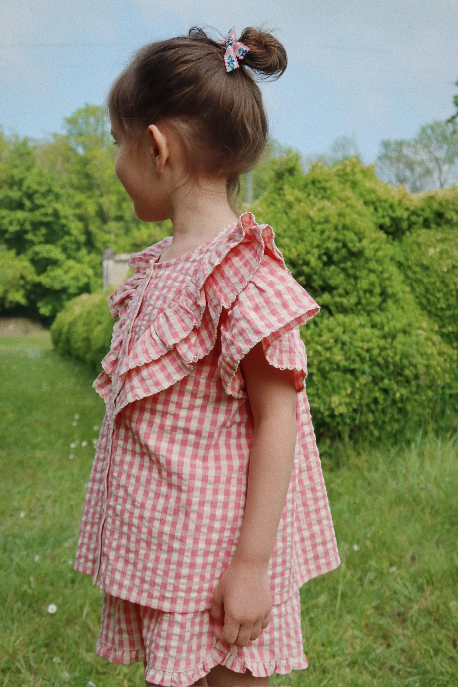 A young child with brown hair in a bun stands outdoors on grass, wearing Konges Slojd Girls' Kim Frill Shorts in pink and white gingham. Green bushes and trees form the background as she faces to the side.