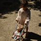 A young child in a light-colored coat pushes a Konges Slojd Sally Doll stroller with two sweet dolls along a sunlit, sandy path.