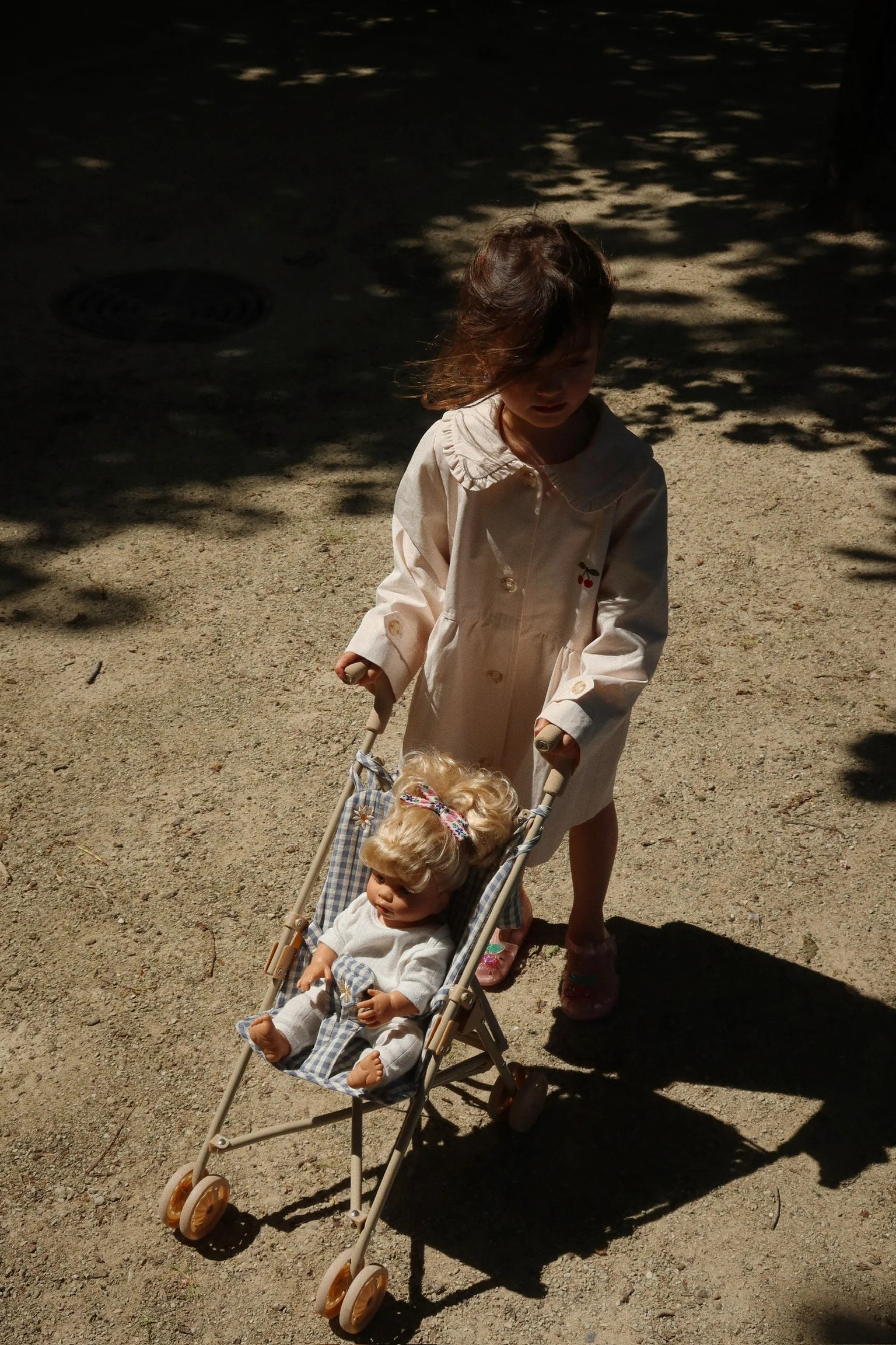 A young child in a light-colored coat pushes a Konges Slojd Sally Doll stroller with two sweet dolls along a sunlit, sandy path.