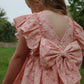 A young girl with light brown hair wears the Konges Slojd Girls' Ava Bow Dress, featuring frill details and a large back bow, while standing outdoors on the grass.