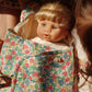 The Konges Slojd Sally Doll, with blonde hair and blue eyes, lies wrapped in a colorful floral blanket, surrounded by partially visible children.