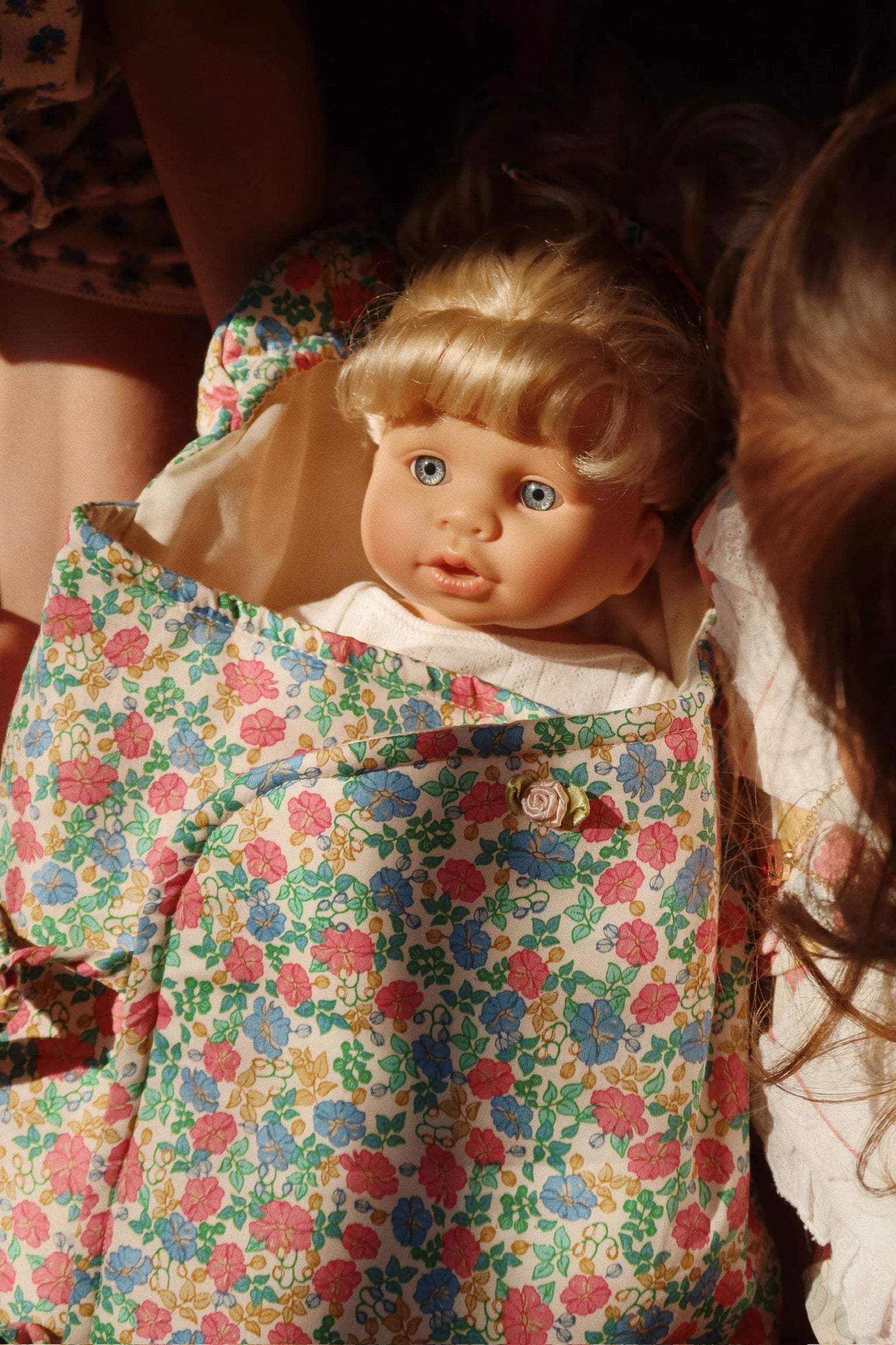 The Konges Slojd Sally Doll, with blonde hair and blue eyes, lies wrapped in a colorful floral blanket, surrounded by partially visible children.
