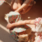 Two children in floral-patterned clothes sit on the floor, wrapping the Konges Slojd Sally Doll in a floral blanket as sunlight casts soft shadows over the cozy scene.