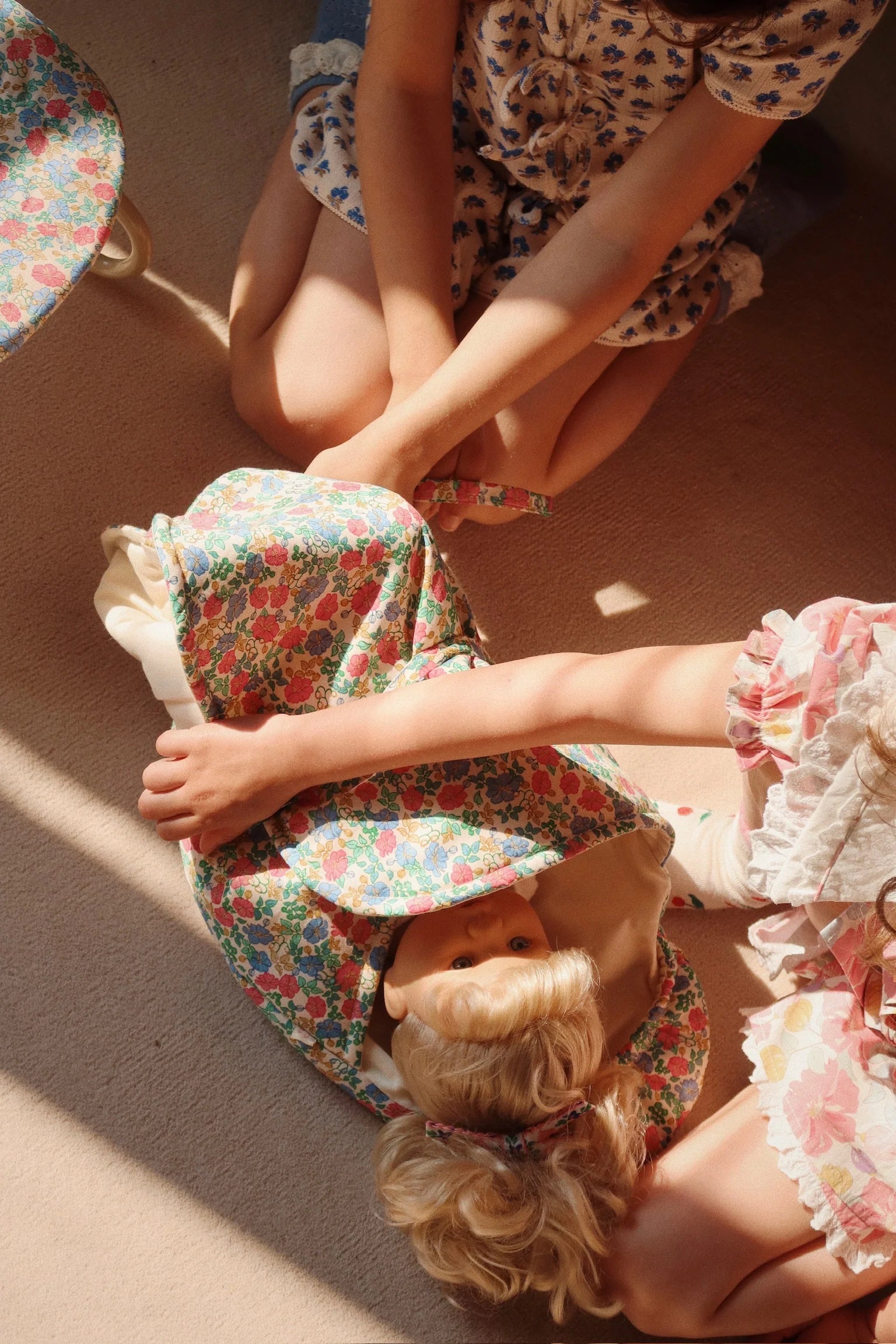 Two children in floral-patterned clothes sit on the floor, wrapping the Konges Slojd Sally Doll in a floral blanket as sunlight casts soft shadows over the cozy scene.