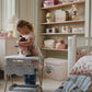 A young girl plays with the Konges Slojd Doll Bouncer on a cotton cushion seat in a pastel-themed nursery, while shelves behind her hold toys, boxes, and decor in soft colors.