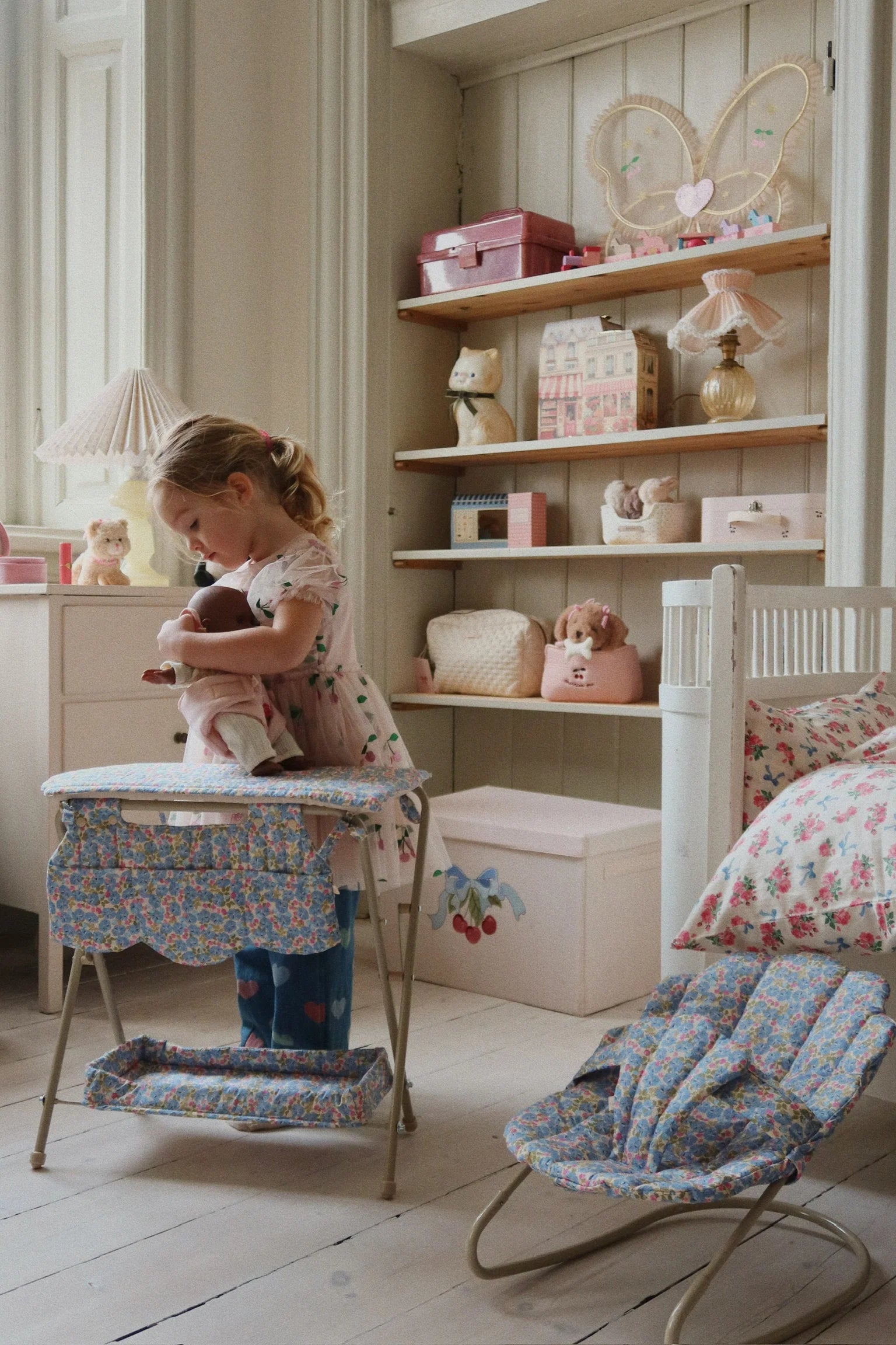 A young girl plays with the Konges Slojd Doll Bouncer on a cotton cushion seat in a pastel-themed nursery, while shelves behind her hold toys, boxes, and decor in soft colors.
