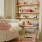 A tidy nursery with a white crib, toy-filled shelves, a baby doll on the table, and the Konges Slojd Doll Changing Table—a pink cradle with storage pockets and metal frame—placed on the wooden floor.