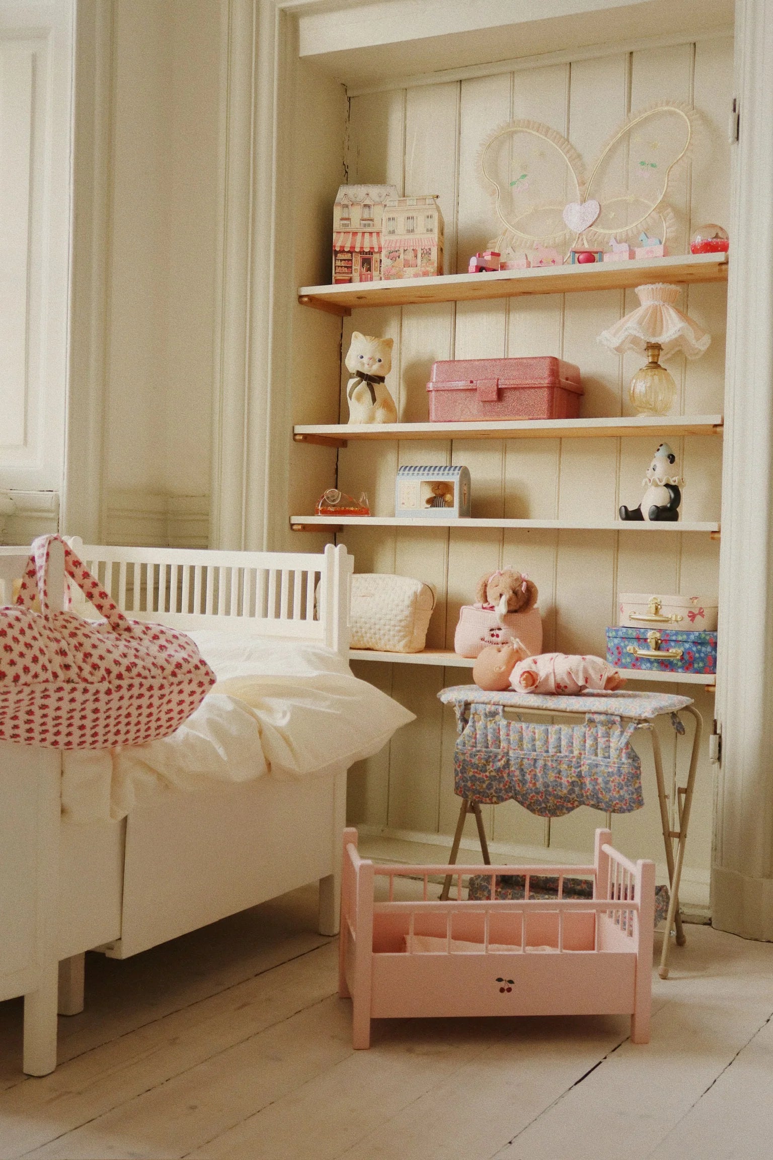 A tidy nursery with a white crib, toy-filled shelves, a baby doll on the table, and the Konges Slojd Doll Changing Table—a pink cradle with storage pockets and metal frame—placed on the wooden floor.