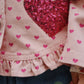 Close-up of a child’s outfit featuring Konges Slojd’s Baby Girls' and Girls' Nola Sequin Shirt Jacket with pink cotton twill, heart prints, sequins, frill hems, a large red sequin heart, and blue jeans with a green heart print.