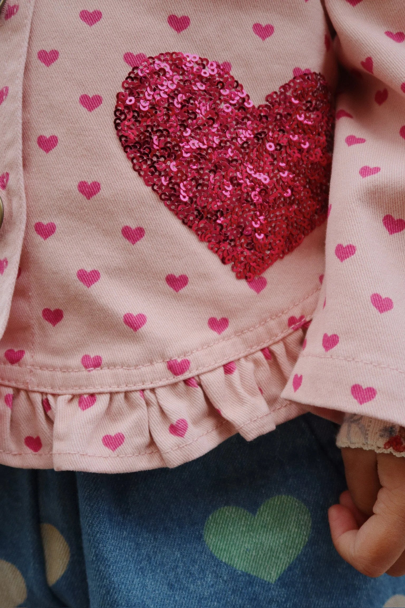 Close-up of a child’s outfit featuring Konges Slojd’s Baby Girls' and Girls' Nola Sequin Shirt Jacket with pink cotton twill, heart prints, sequins, frill hems, a large red sequin heart, and blue jeans with a green heart print.