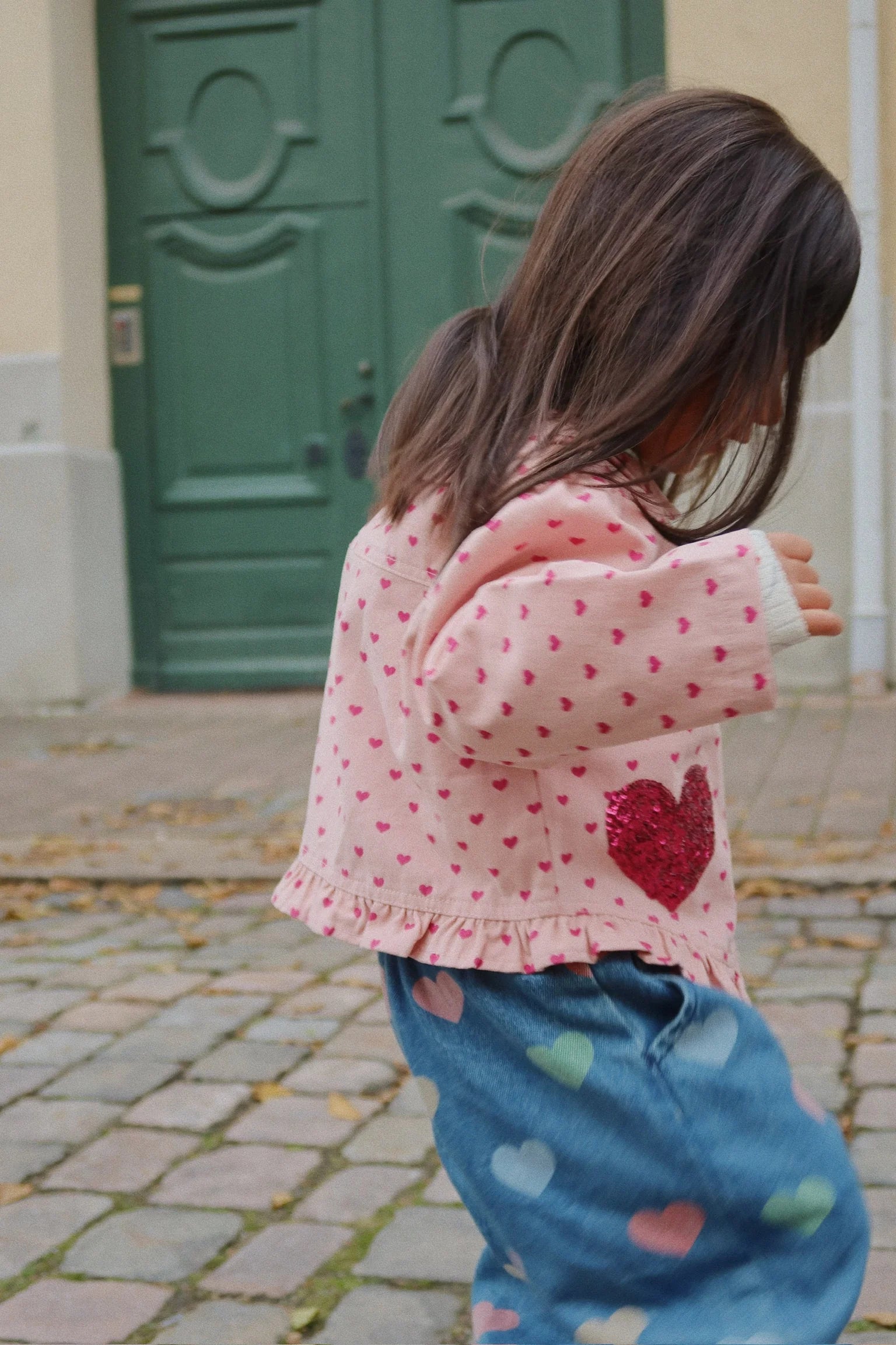 A young girl walks outside on a cobblestone path, wearing the Konges Slojd Baby Girls' and Girls' Nola Sequin Shirt Jacket.