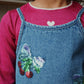 A child in the Konges Slojd Baby Girls' and Girls' Roli Blouse in pink with a heart detail at the collar, paired with a denim dress featuring sequin cherries, a bow, and shimmering lurex thread.