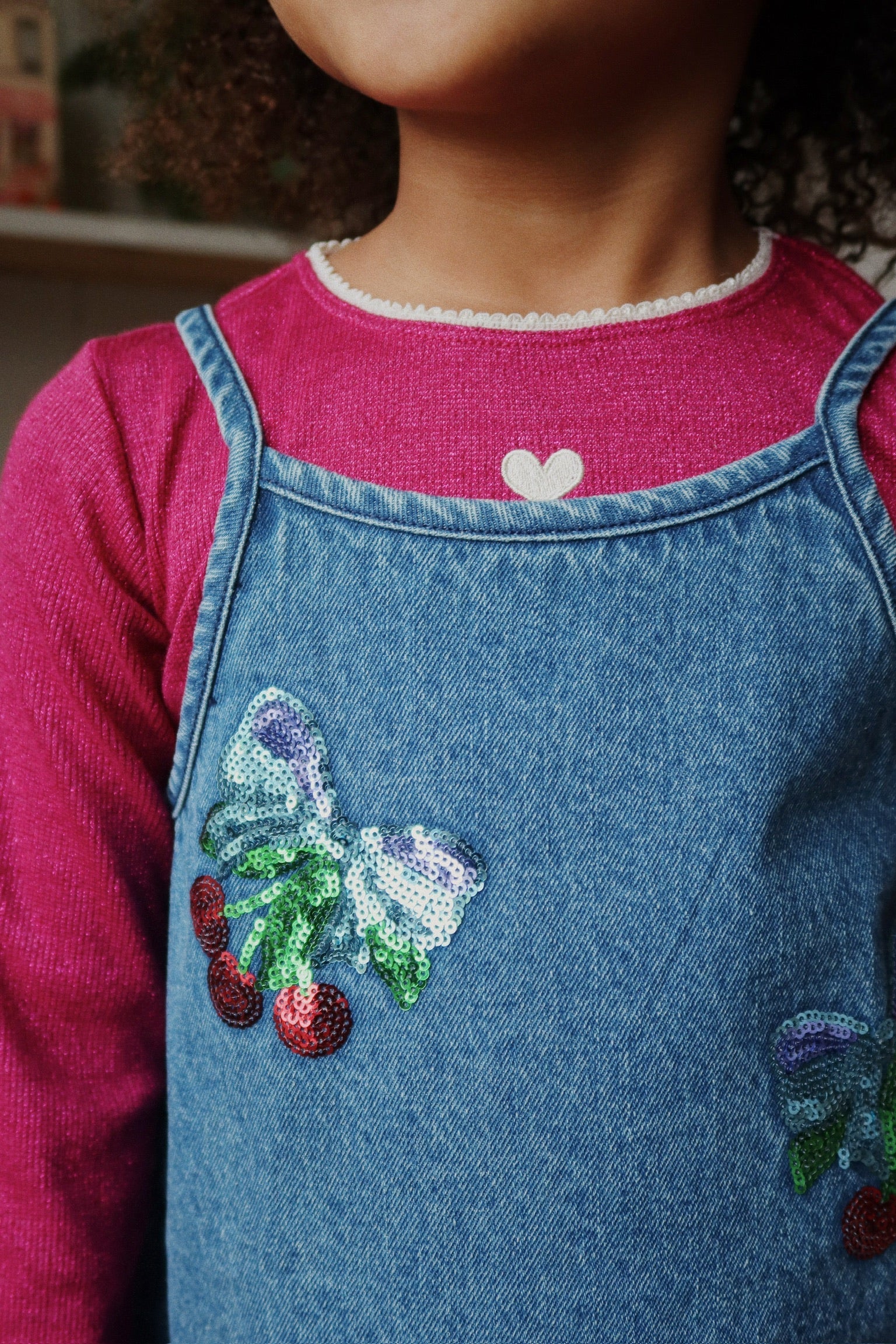 A child in the Konges Slojd Baby Girls' and Girls' Roli Blouse in pink with a heart detail at the collar, paired with a denim dress featuring sequin cherries, a bow, and shimmering lurex thread.