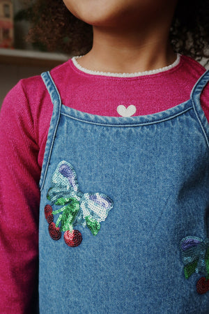 A child in the Konges Slojd Baby Girls' and Girls' Roli Blouse in pink with a heart detail at the collar, paired with a denim dress featuring sequin cherries, a bow, and shimmering lurex thread.