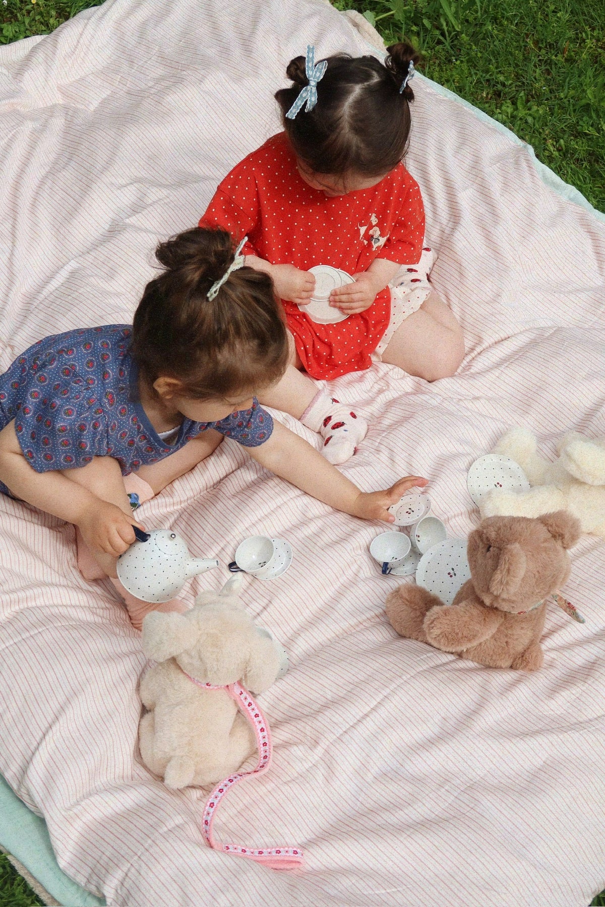 Two young children sit on a striped blanket, using a Konges Slojd Blue Dot Tea Party Set to host a pretend play session for their two stuffed animals.