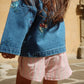 A child with long brown hair wears the Konges Slojd Girls’ Magot Sequin Denim Jacket, paired with pink striped shorts, while standing outdoors on a stone surface.