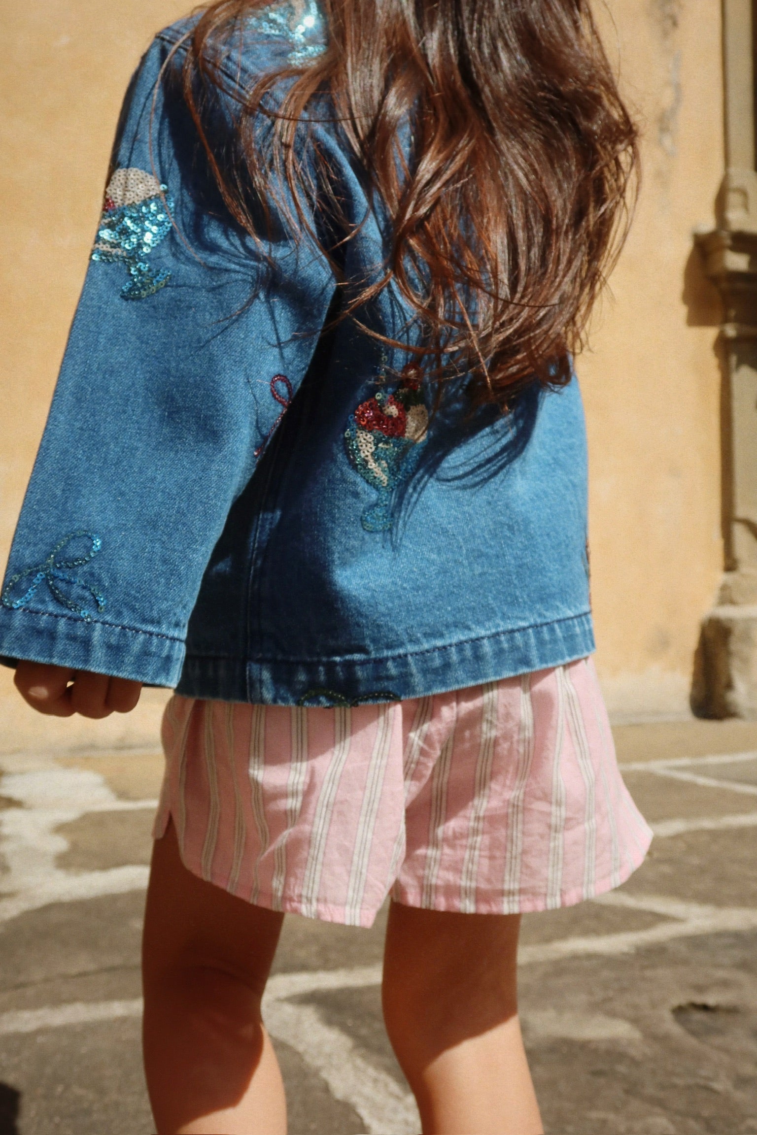 A child with long brown hair wears the Konges Slojd Girls’ Magot Sequin Denim Jacket, paired with pink striped shorts, while standing outdoors on a stone surface.