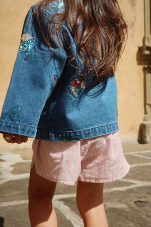 A child with long brown hair wears the Konges Slojd Girls’ Magot Sequin Denim Jacket, paired with pink striped shorts, while standing outdoors on a stone surface.