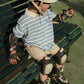 A young child wearing a helmet, elbow pads, knee pads, rollerblades, and the Konges Slojd Boy’s Spotty Short Sleeve Polo sits on a green bench outdoors.