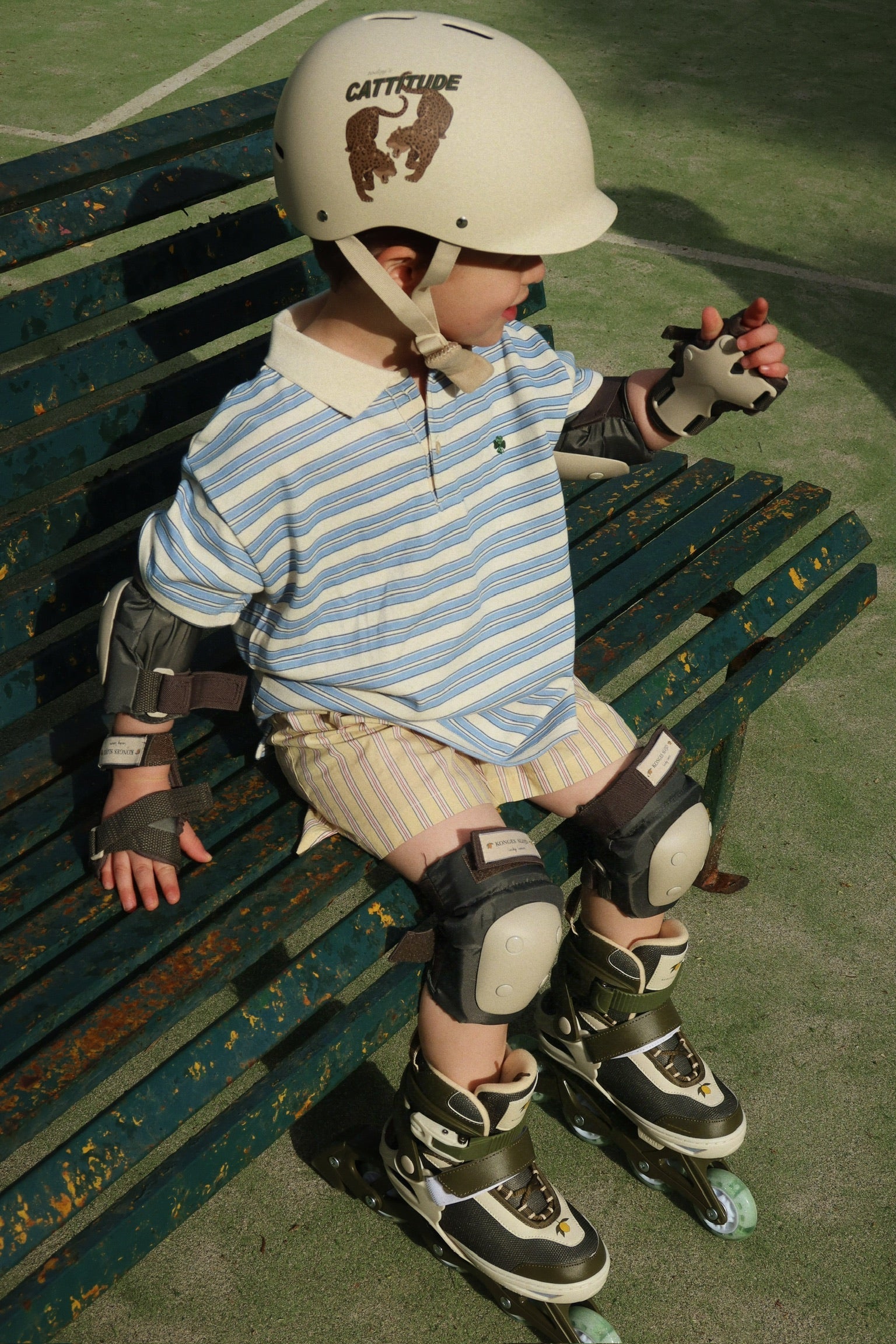 A young child wearing a helmet, elbow pads, knee pads, rollerblades, and the Konges Slojd Boy’s Spotty Short Sleeve Polo sits on a green bench outdoors.