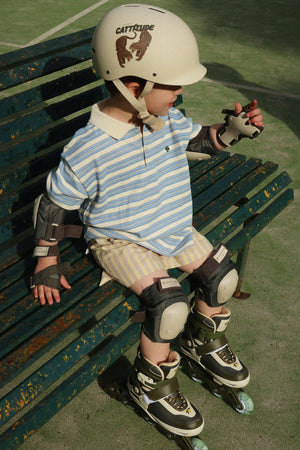 A young child wearing a helmet, elbow pads, knee pads, rollerblades, and the Konges Slojd Boy’s Spotty Short Sleeve Polo sits on a green bench outdoors.