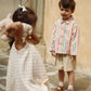 A little girl holding a plush toy stands in front of a boy wearing the Konges Slojd Baby Boys’ and Boys’ Franzen Linen Shorts, paired with a striped shirt and sandals, outdoors by a stone wall.
