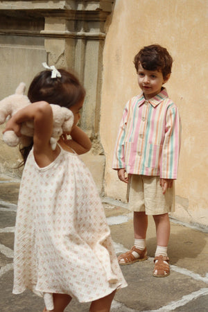 A little girl holding a plush toy stands in front of a boy wearing the Konges Slojd Baby Boys’ and Boys’ Franzen Linen Shorts, paired with a striped shirt and sandals, outdoors by a stone wall.