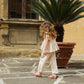 A young child in light pajamas and sandals walks on a stone patio near a potted palm, with a tall window and yellow wall in the background. She wears the Konges Slojd Baby Girls’ and Girls’ Coco Top, crafted from soft muslin—perfect for keeping cool on warm days.