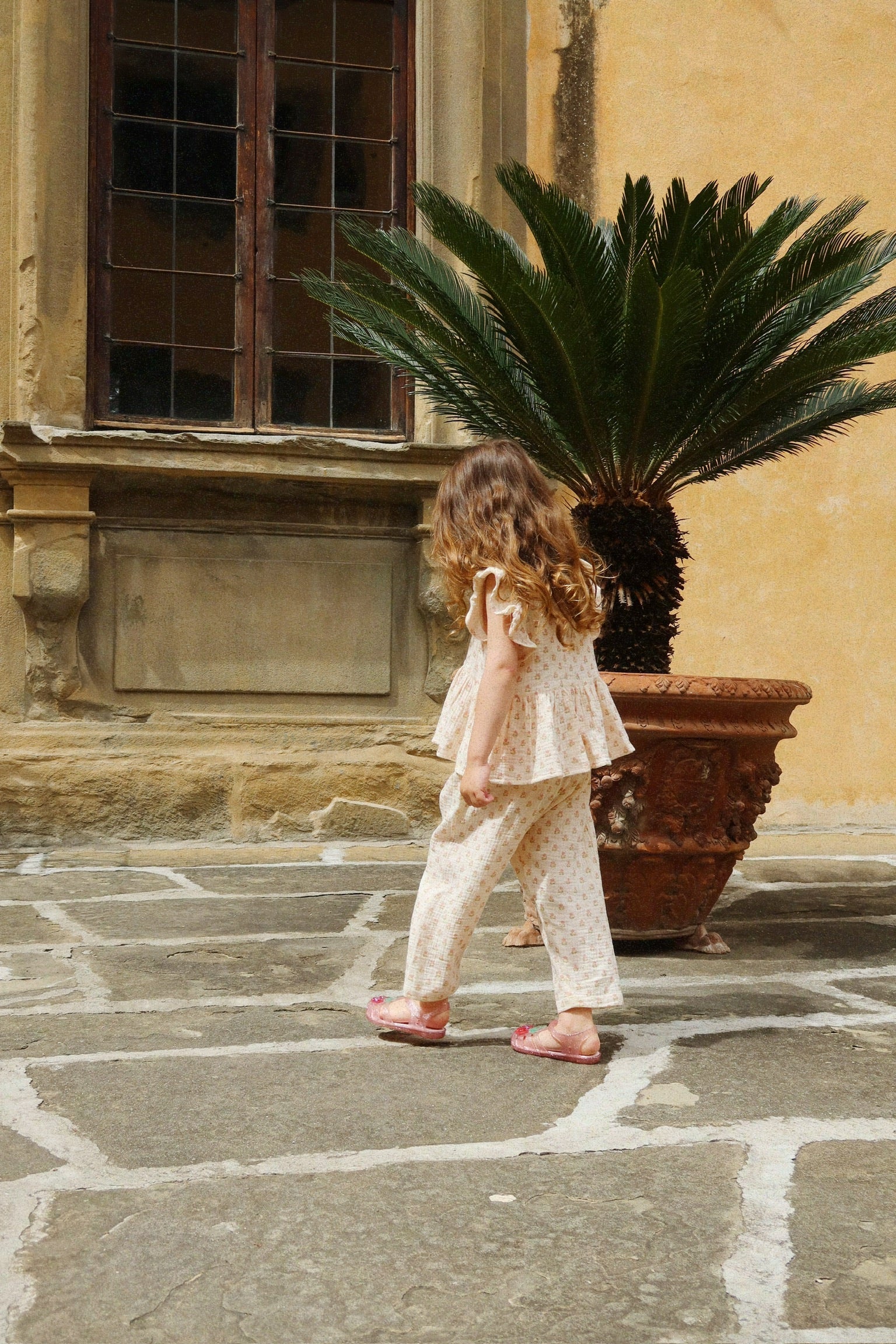 A young child in light pajamas and sandals walks on a stone patio near a potted palm, with a tall window and yellow wall in the background. She wears the Konges Slojd Baby Girls’ and Girls’ Coco Top, crafted from soft muslin—perfect for keeping cool on warm days.