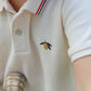 Close-up of a child wearing the Konges Slojd Baby Boys’ and Boys’ Florian Polo Shirt, a white polo featuring a classic button placket and regular collar detailed with red and blue stripes, plus a small embroidered lemon accent, holding the worn handle of a tennis racket.