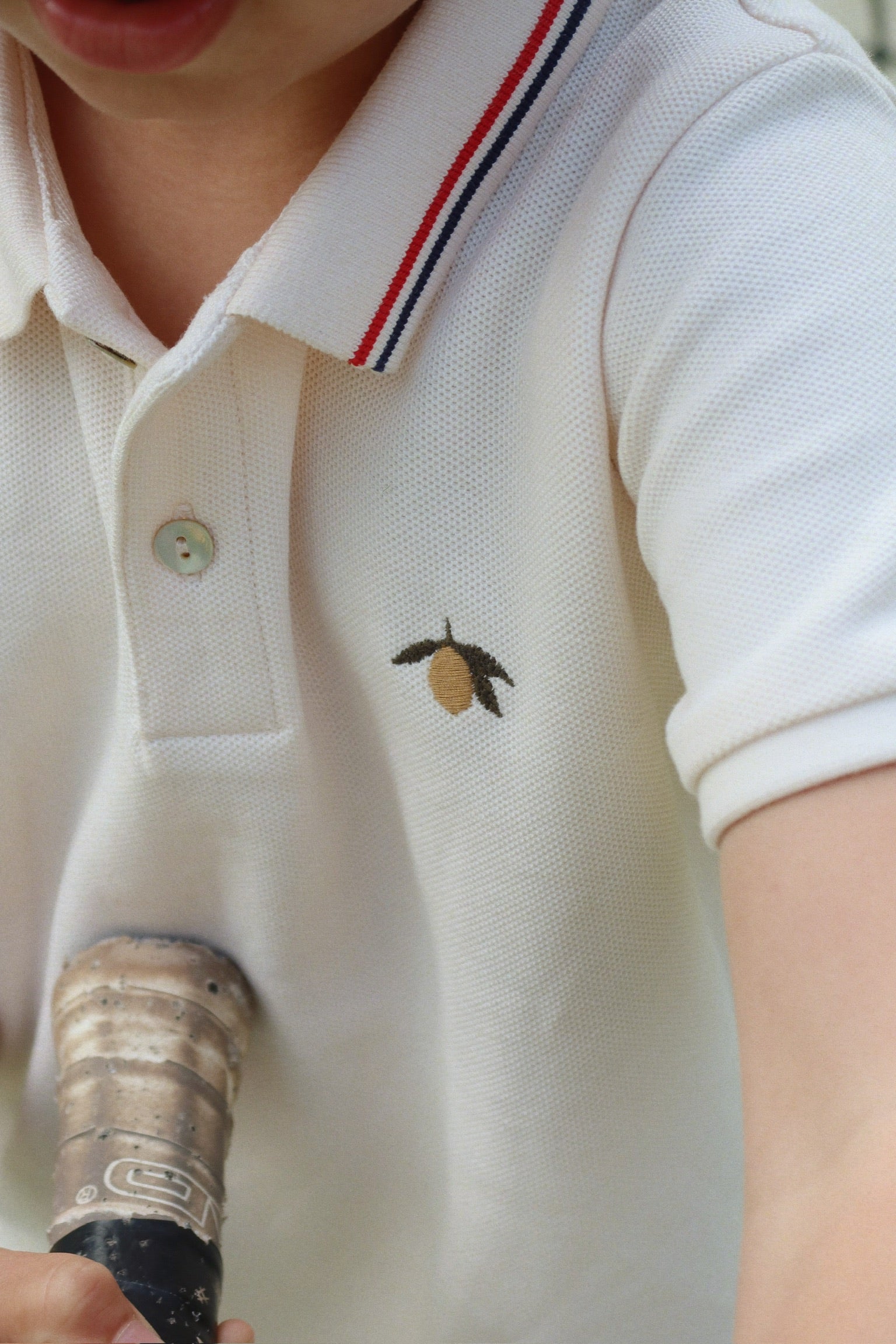 Close-up of a child wearing the Konges Slojd Baby Boys’ and Boys’ Florian Polo Shirt, a white polo featuring a classic button placket and regular collar detailed with red and blue stripes, plus a small embroidered lemon accent, holding the worn handle of a tennis racket.