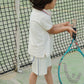 A young child stands by the net on an outdoor tennis court, holding a racket with both hands and dressed in Konges Slojd Baby Boys’ and Boys’ Florian Shorts—crafted from organic cotton and featuring a comfortable drawstring waist.