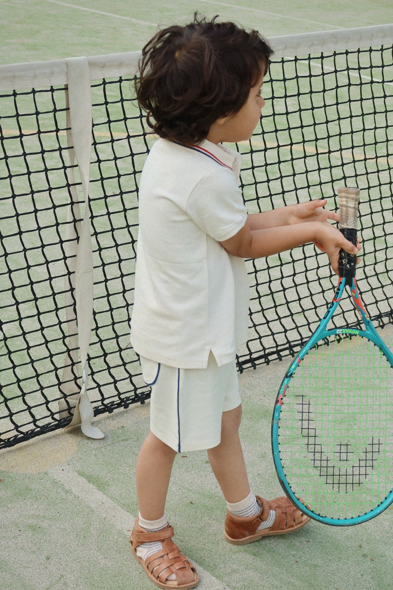 A young child stands by the net on an outdoor tennis court, holding a racket with both hands and dressed in Konges Slojd Baby Boys’ and Boys’ Florian Shorts—crafted from organic cotton and featuring a comfortable drawstring waist.