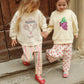 Two young children wearing Konges Slojd Baby Girls’ and Girls’ Frankie Pants and cream sweatshirts walk down stone steps while holding hands, in front of a wooden door.
