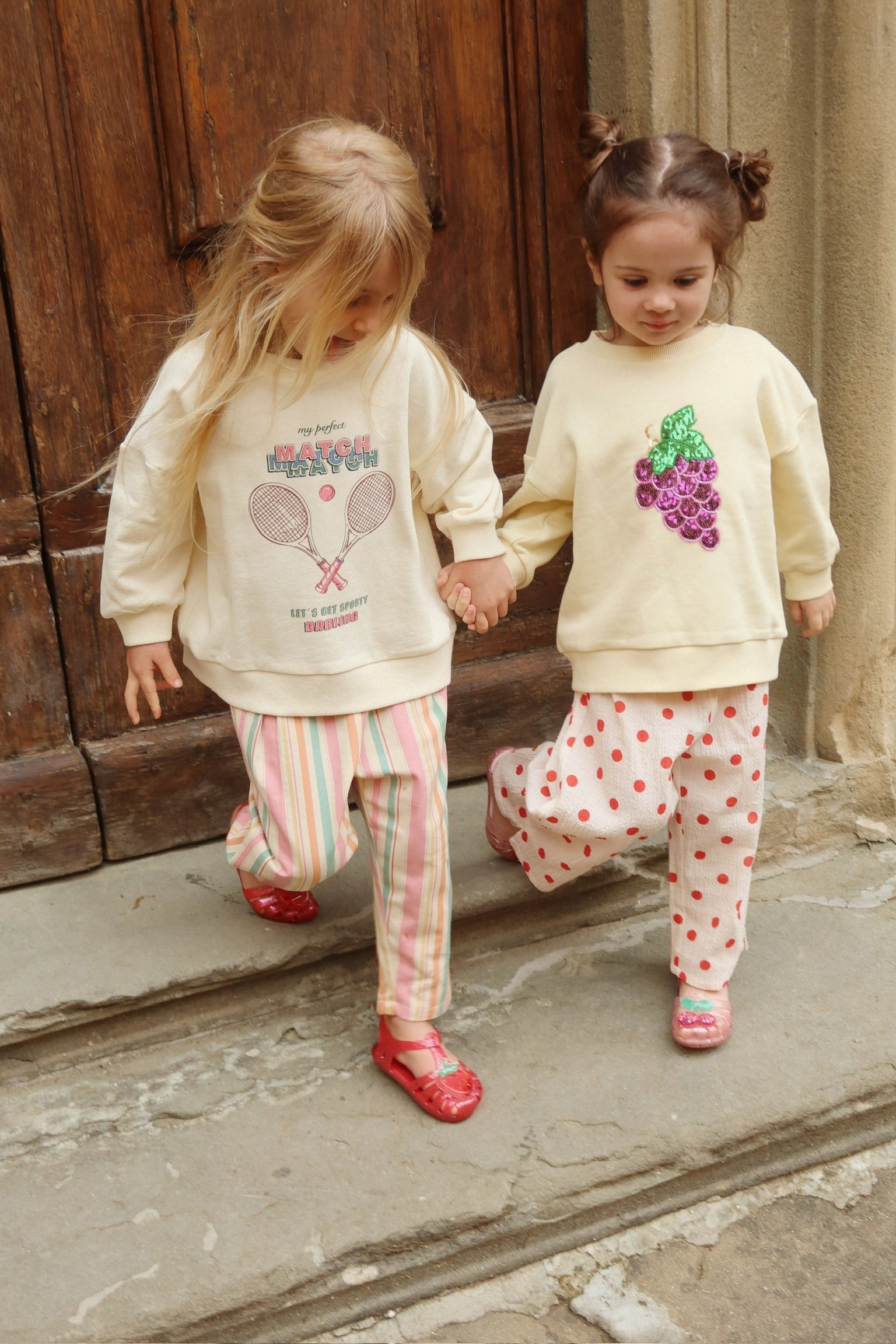 Two young children wearing Konges Slojd Baby Girls’ and Girls’ Frankie Pants and cream sweatshirts walk down stone steps while holding hands, in front of a wooden door.