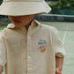 A young child wearing the Konges Slojd Baby Boys’ and Boys’ Franzen Short Sleeve Linen Shirt, featuring a “Sour and Salty” fish and lemon design with a matching hat, stands on a tennis court.