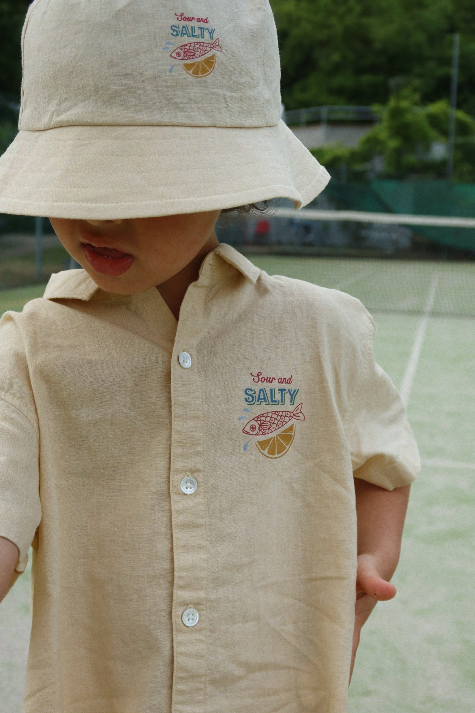 A young child wearing the Konges Slojd Baby Boys’ and Boys’ Franzen Short Sleeve Linen Shirt, featuring a “Sour and Salty” fish and lemon design with a matching hat, stands on a tennis court.
