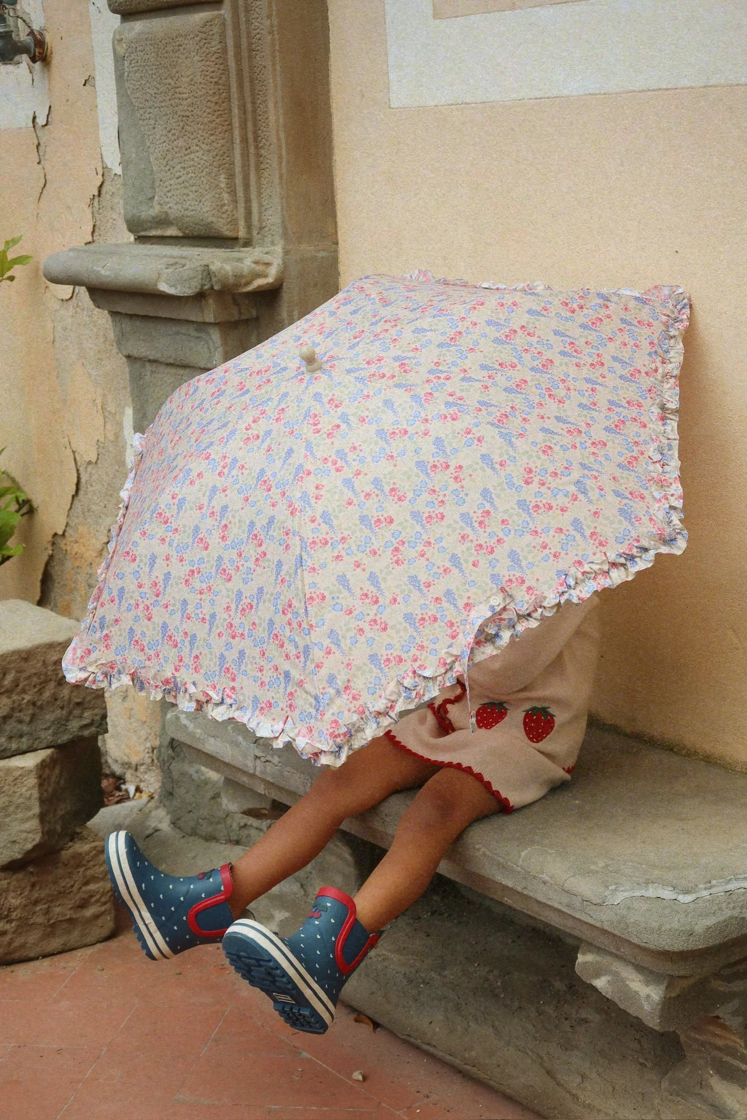 A child sits on a stone bench, face and upper body hidden by a floral-patterned umbrella, wearing blue sneakers and the Konges Slojd Girls' Collette Dress.