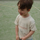A young boy stands on a tennis court, smiling, wearing the Konges Slojd Baby Boys’ Itty Tee and blue striped shorts.