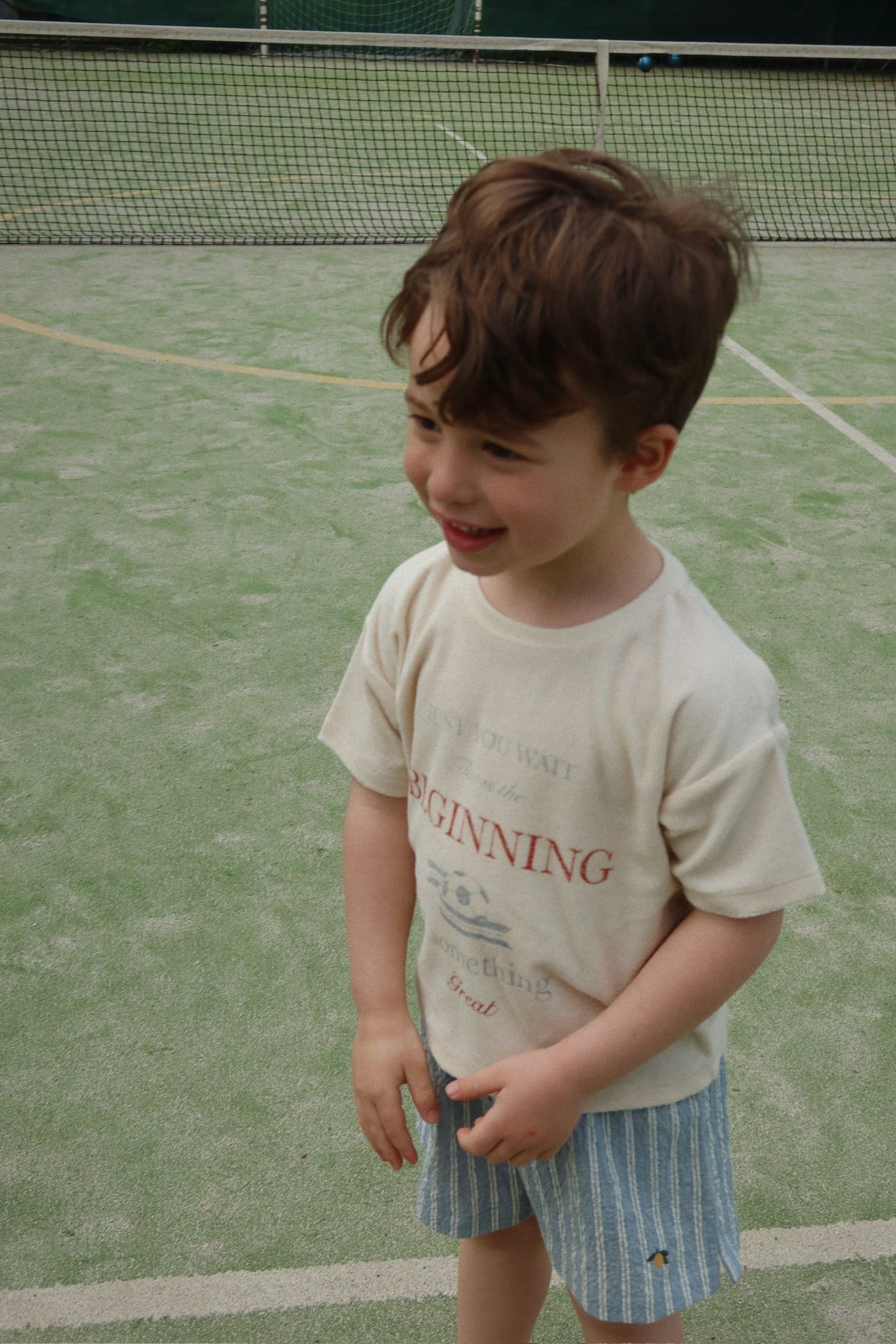 A young boy stands on a tennis court, smiling, wearing the Konges Slojd Baby Boys’ Itty Tee and blue striped shorts.