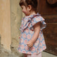 A toddler with brown hair styled in a bun, wearing the Konges Slojd Baby Girls’ and Girls’ Kim Frill Shirt, stands smiling sideways next to a wooden door and textured wall.