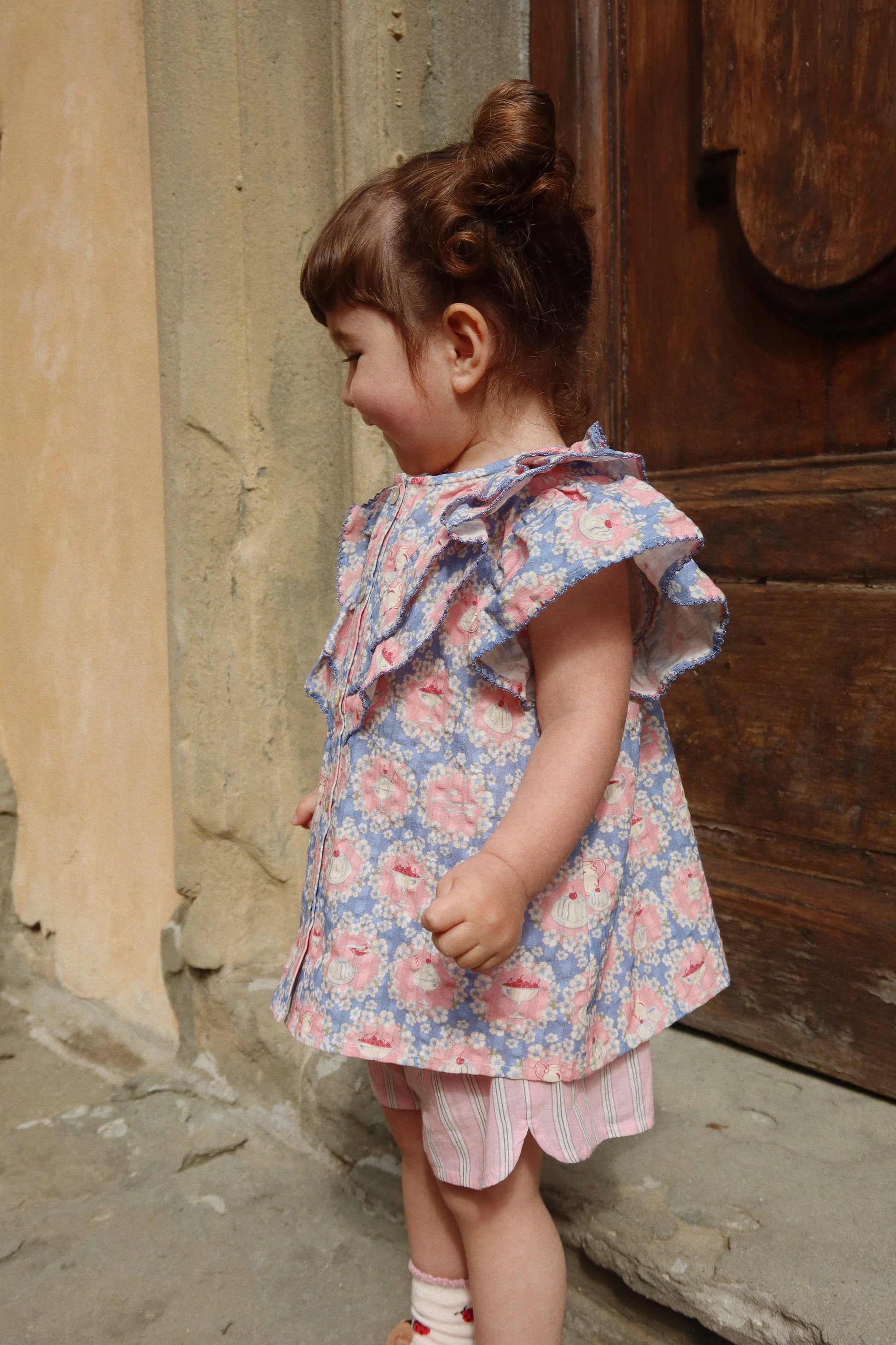 A toddler with brown hair styled in a bun, wearing the Konges Slojd Baby Girls’ and Girls’ Kim Frill Shirt, stands smiling sideways next to a wooden door and textured wall.