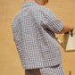 A young child stands sideways against a tan wall, holding a watering can and wearing the Konges Slojd Baby Boys’ and Boys’ Kim Short Sleeve Shirt in a classic blue and white checkered seersucker.